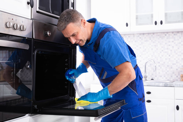 Close-up Of A Smiling Male Janitor Cleaning Oven With Yellow Napkin In The Kitchen - Photo, Image