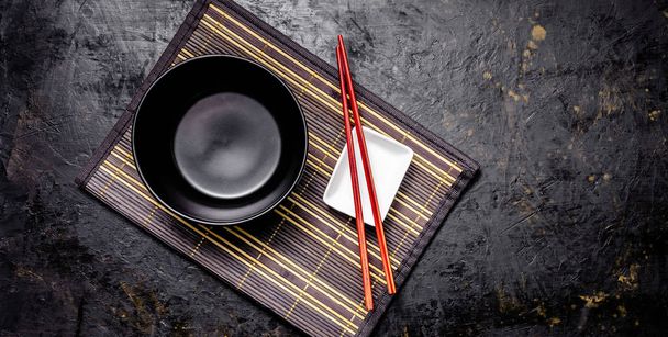 Empty Japanese dishes. A black ceramic bowl for Chinese noodles or Thai soup lies on a bamkuk rug. White saucepot for soy sauce and red Chinese sticks on a black background. Top view, copy space - Foto, immagini