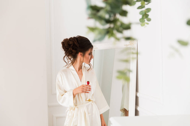 The beautiful young bride standing in front of the mirror and holding a glass of champagne with strawberry in hand indoor - Foto, afbeelding