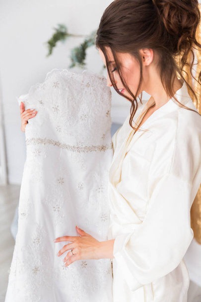 Side view of the beautiful young bride who is standing indoor and holding her bridal gown and looking at it - Fotó, kép