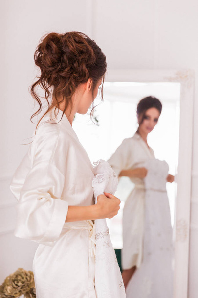 Kaunis nuori morsian seisoo peilin edessä sisätiloissa ja pitää morsiuspukuaan
 - Valokuva, kuva