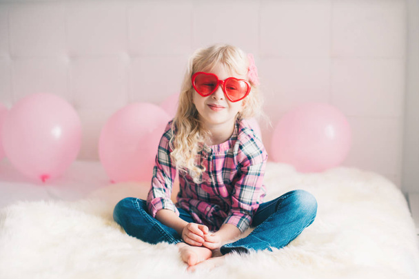 Portré boldog fehér kaukázusi aranyos aranyos vicces lány szív alakú szemüveget visel. Gyermek hálószoba ünneplése Valentin nap ünnep ágyon ül.  - Fotó, kép