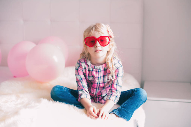 Portrait of happy white Caucasian cute adorable funny girl wearing heart shape glasses. Child sitting on bed in bedroom celebrating Valentine Day holiday.  - Foto, Bild