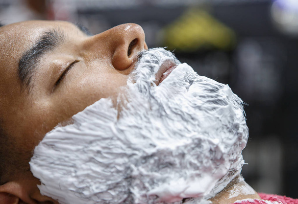 Porträt eines jungen schwarzen Mannes mit weißer Rasiercreme im Gesicht in barbershop.clients Gesicht mit Schaum für genaue Rasur im Salon.male Schönheitsbehandlungskonzept bedeckt - Foto, Bild