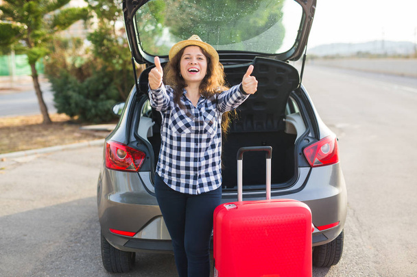 Ταξίδια, διακοπές και άνθρωποι έννοια - νεαρή γυναίκα στέκεται κοντά στο πίσω μέρος του αυτοκινήτου χαμογελώντας και ετοιμάζεται να πάει, κρατώντας το καπέλο - Φωτογραφία, εικόνα