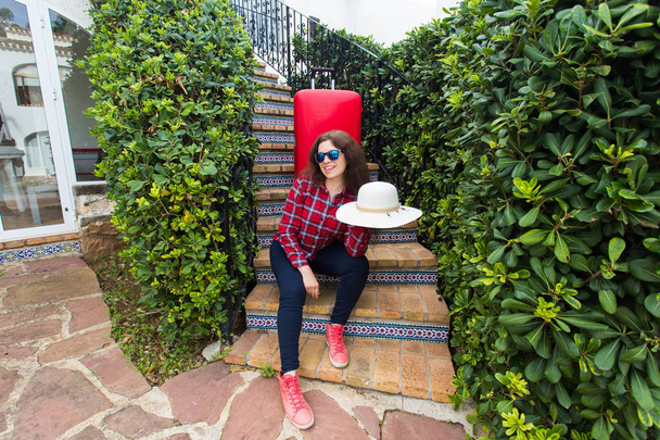 Cestování, dovolených a svátků koncept - happy cestovatelů žena sedí na schodech v sluneční brýle s červenými kufr a čepice - Fotografie, Obrázek