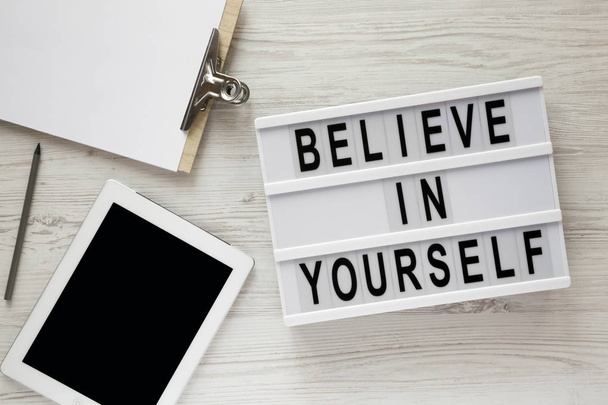 Modern board with text 'Believe in yourself', tablet, clipboard and sheet of paper on a white wooden surface, top view. Overhead. - Фото, изображение