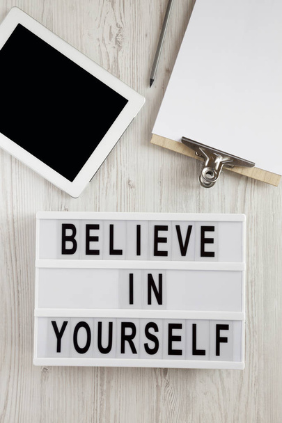 moderne Tafel mit Text "Glaube an dich selbst", Tablet, Klemmbrett und Blatt Papier auf weißem Holzgrund, Draufsicht. - Foto, Bild