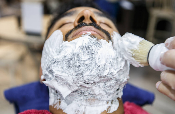 Barber spreads white shaving foam on client face with professional swob brush in barbershop salon.Hairdresser spreading cream on young black man beard before shave.Male beauty treatment concept - Fotó, kép