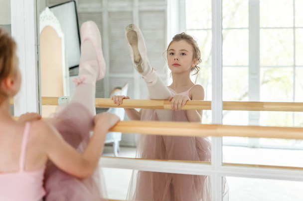 Młoda tancerka klasyczna w klasie tańca. Piękne, pełne wdzięku baletnice ćwiczą pozycje baletowe w różowej spódnicy tutu w pobliżu dużego lustra w białej hali oświetleniowej - Zdjęcie, obraz