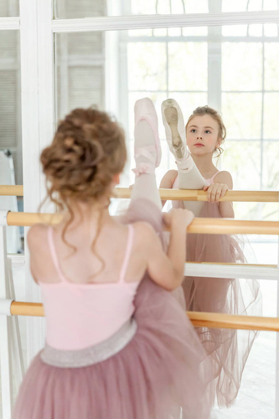 Молодая танцовщица балета в танцевальном классе. Красивая изящная балерина практикует балетные позиции в розовой юбке в пачке возле большого зеркала в белом светлом зале - Фото, изображение