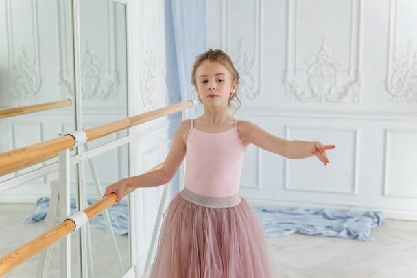 Молодая танцовщица балета в танцевальном классе. Красивая изящная балерина практикует балетные позиции в розовой юбке в пачке возле большого зеркала в белом светлом зале - Фото, изображение