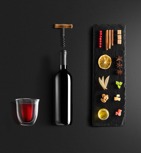 Mulled wine recipe ingredients and kitchen accessories, bottle of red wine, cinnamon, anise stars, orange, brown sugarand spice on black background. - Photo, image