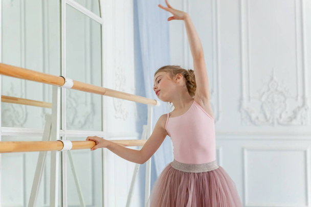 Een jong klassiek balletdanseres meisje in dansles. Prachtige sierlijke ballerina praktijk ballet posities in roze tutu rok in de buurt van grote spiegel in wit licht hal - Foto, afbeelding