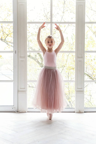 Een jong klassiek balletdanseres meisje in dansles. Prachtige sierlijke ballerina praktijk ballet posities in roze tutu rok in de buurt van groot raam in wit licht hal - Foto, afbeelding