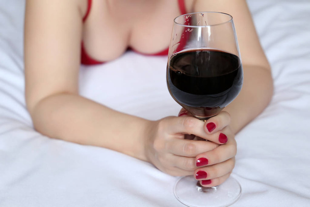 Woman drinking wine in the bed, romantic date. Sexy girl in lingerie lying with a glass of red wine in her hands, concept of relaxation, sex party, drunk woman, female alcoholism - Photo, image