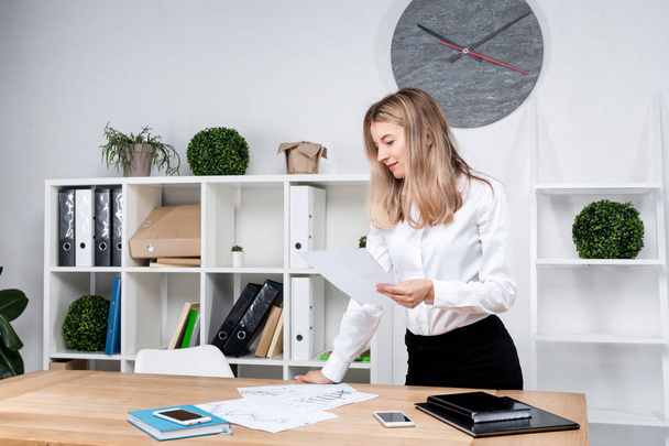 Theme business woman at work. Beautiful young caucasian woman business man working standing in the office near the table, checks edits financial charts on paper. - Foto, immagini