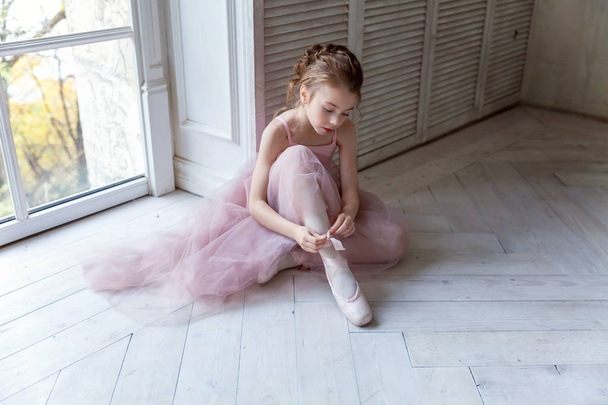 Joven bailarina de ballet clásica en clase de baile. Hermosa bailarina elegante en falda de tutú rosa se pone zapatos puntiagudos cerca de una gran ventana en salón de luz blanca - Foto, Imagen