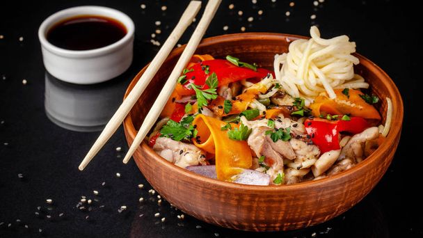 Japanese or Chinese udon noodles with chicken and vegetables, balkar pepper, carrots, parsley, white and black sesame, red onion. noodles in a clay red plate, next to soy sauce and sticks - Foto, afbeelding