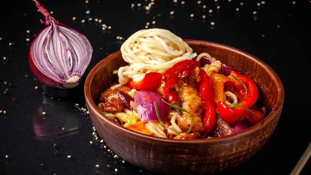Japanese or Chinese udon noodles with chicken and vegetables, balkar pepper, carrots, parsley, white and black sesame, red onion. noodles in a clay red plate, next to soy sauce and sticks - Photo, Image