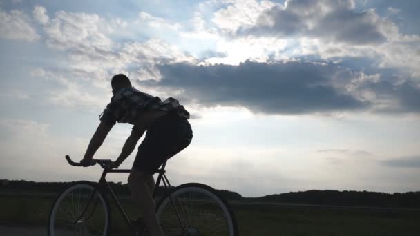 Silueta mladík na koni na vintage kolo s krásného západu slunce na obloze v pozadí. Chlap na kole v zemi silnici. Mužské cyklistu jedoucího pevné ozubené kolo. Zdravý aktivní životní styl zpomalené - Záběry, video