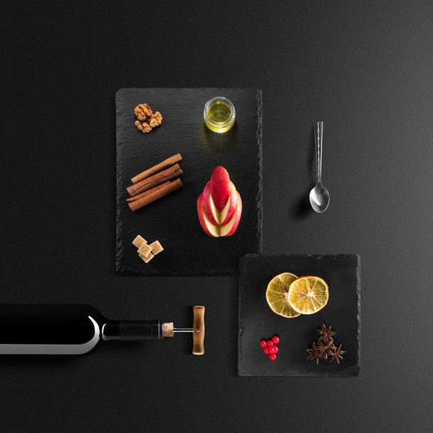 Mulled wine recipe ingredients and kitchen accessories, bottle of red wine, cinnamon, anise stars, orange, brown sugarand spice on black background. - Photo, Image
