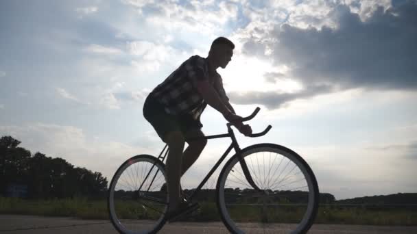 Silhouette of young man riding at vintage bicycle with beautiful sunset sky at background. Sporty guy cycling in the country road. Male cyclist riding bike. Healthy active lifestyle Slow motion - Footage, Video