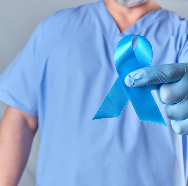 Doktor üniformalı ve lateks eldivenli elinde mavi bir kurdele var. Prostat kanserinin tedavi ve savaşının sembolü. - Fotoğraf, Görsel