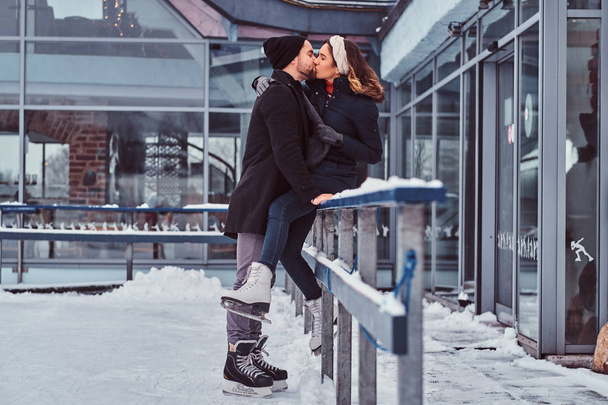 Ελκυστικό ζευγάρι στην αγάπη, ημερομηνία στο παγοδρόμιο, ένα κορίτσι που κάθεται σε ένα προστατευτικό κιγκλίδωμα και το φιλί με το φίλο της. - Φωτογραφία, εικόνα