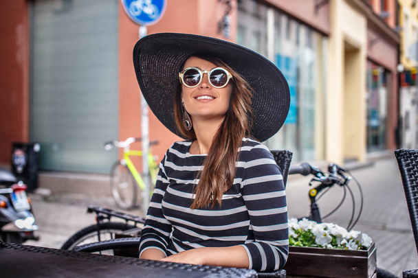 Tyylikkäästi pukeutunut nuori nainen yllään tyylikäs hattu ja aurinkolasit istuu kesällä katu kahvila
. - Valokuva, kuva