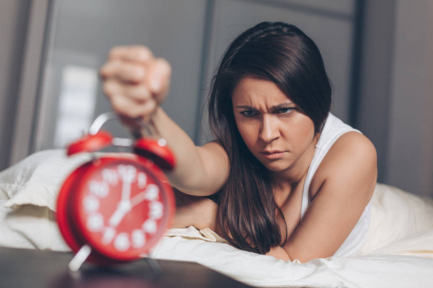 Angry young woman kill off alarm clock by fist on the bed in the morning. Awakening of the sleeping woman. Focus on woman. - Fotografie, Obrázek