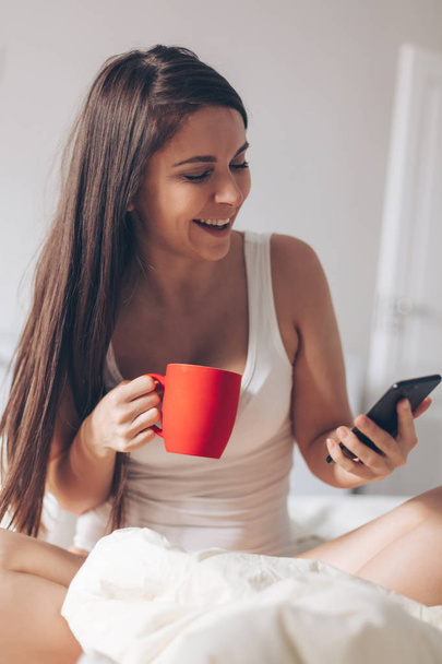 Young smiling woman drinking coffee at home in her bed and checking her smartphone - Фото, изображение