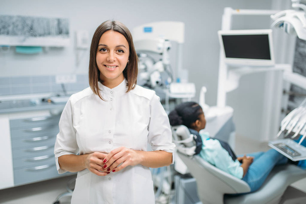Glimlachend vrouwelijk tandarts, tandheelkundige kliniek, patiënt in de stoel op de achtergrond. Vrouw in de tandheelkunde kabinet stomatologie, tanden zorg - Foto, afbeelding