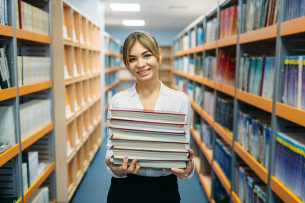 Vrij vrouwelijke persoon met stapel boeken tussen de schappen in de Universiteitsbibliotheek. Jonge vrouw in de leeszaal, kennis depositaris - Foto, afbeelding
