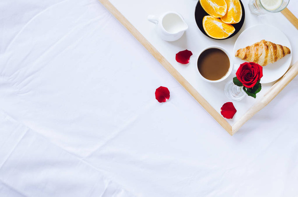 Ρομαντικό ημέρας βαλεντίνων πρωινό στο κρεβάτι, δίσκος με φρέσκα κρουασάν, καφέ εσπρέσο με γάλα, φρέσκα πορτοκάλια και κόκκινο τριαντάφυλλο. Καλημέρα έννοια. Απολαύστε την αργή ζωή. Το top view. Χώρο αντίγραφο. - Φωτογραφία, εικόνα
