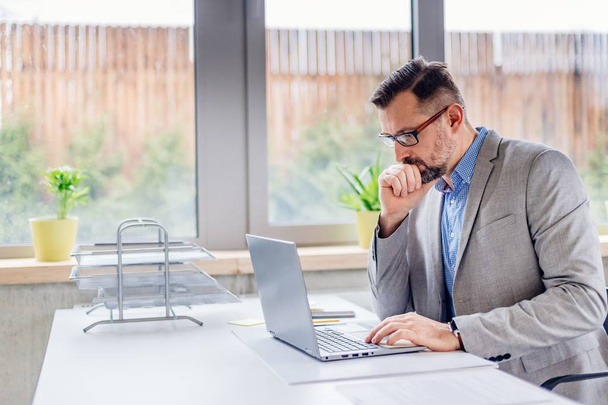 Promyšlené středního věku pohledný podnikatel v košili pracují na přenosném počítači v kanceláři. Muži, kteří pracují v kanceláři - Fotografie, Obrázek