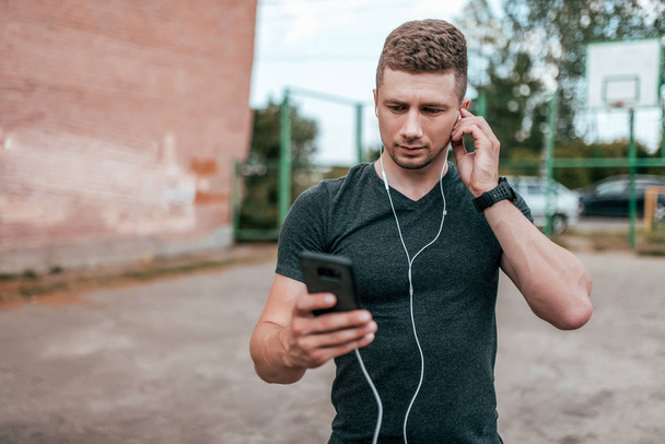 Young guy athlete man, serious look screen of smartphone, talking on video call. Summer in city park on background of sports field. In casual clothes with headphones, listening to music. - Фото, зображення
