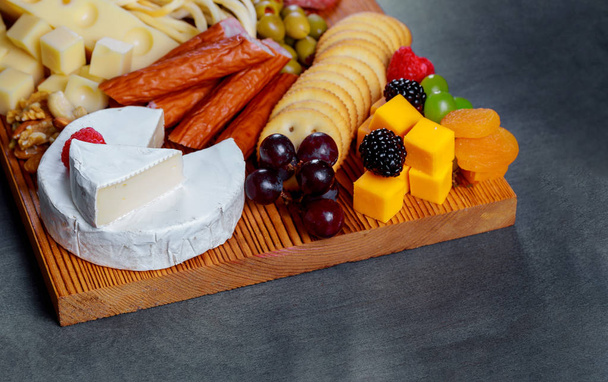 Cutting wood board with fresh assorted cheese and meat crackers, green olives, nuts and berries on gray boards - Photo, Image