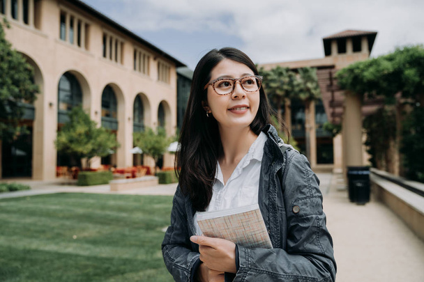 Attractive smiling charming asian young girl student with school book relaxing walking in university outdoor. college lady enjoy sunshine at the park in stanford surrounding by green grass and trees. - Φωτογραφία, εικόνα