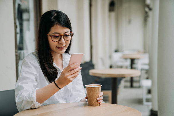 Vonzó ázsiai nő, egy utcai kávézó szabadtéri asztalnál ül ő intelligens telefonról szöveges üzenetek olvasása. fiatal elegáns office hölgy pihentető délutáni tea időben a munkából. boldog lány szemüveg - Fotó, kép