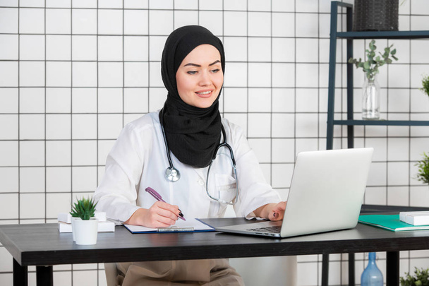Musliminainen lääkäri kirjoittaa raportin työpöydälleen
 - Valokuva, kuva