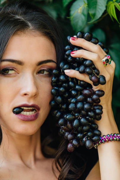 Portrait of young sexy brunette in black dress biting black grapes. - Foto, imagen