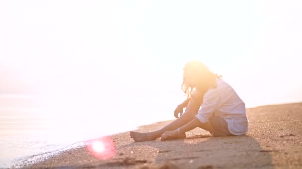 Mulher jovem, bonita, senta-se em uma praia de areia e olha para a distância. Menina feliz sentada à beira-mar esperando por seu namorado
. - Filmagem, Vídeo
