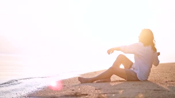 Genç, güzel kadın, kumlu bir plaj üzerinde oturur ve mesafe içine görünüyor. Mutlu kız erkek arkadaşını bekleyen denizin kenarında oturan. - Video, Çekim