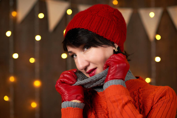 美しい女性は暗い背景の木、クリスマスの照明、フラグ、休日の概念にポーズをとってください。赤のニット帽子とセーターを着てください。. - 写真・画像