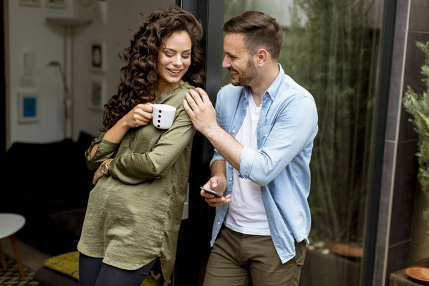 Happy young cute couple in love embracing each other and drinking coffee at home by the window - Photo, Image