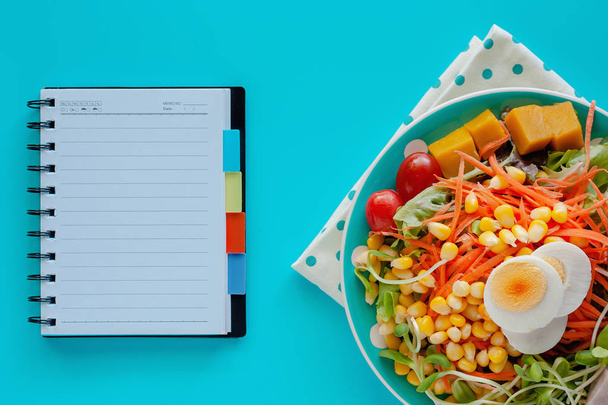Fresh salad vegetable with boiled chicken egg and blank spiral notebook on blue background for healthy eating and diet food concept - Photo, Image