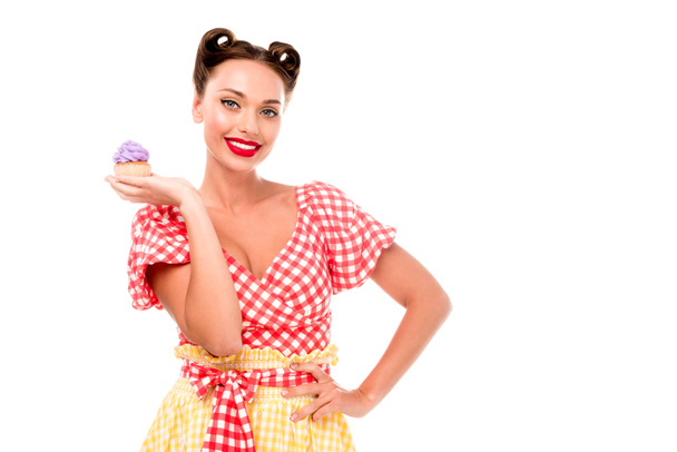 hübsche Frau demonstriert Cupcake mit lila Creme und hält Hand auf Hüfte isoliert auf weiß - Foto, Bild