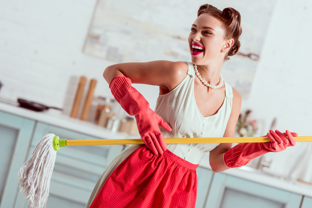 Cheerful pin up girl in red apron and rubber gloves dancing with yellow mop - Φωτογραφία, εικόνα