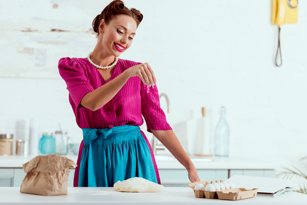 smiling pin up girl in crimson dress and blue apron adding flour to dough  - Photo, Image